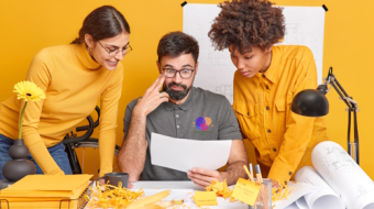 Multicultural partners cooperate on design project discuss ideas look attentively at illustration on paper brainstroming together at blueprint sketches pose in coworking space. Diverse professionals