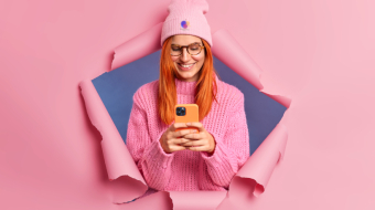 Pretty redhead woman looks happily at smartphone device checks her email smiles pleasantly dressed in stylish clothing poses in knitted jumper through paper hole. Modern life and technology.